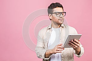 Happy attractive young man standing and using tablet computer isolated over pink background