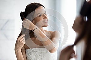 Happy attractive young woman brushing long hair looking in mirror