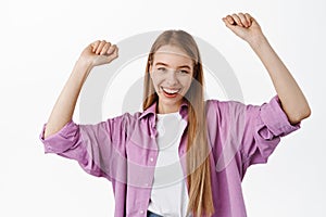 Happy attractive young woman dancing, celebrating and partying, having fun, smiling at camera, raising hands up carefree