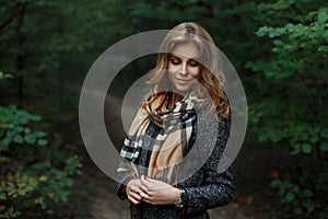 Happy attractive young woman in a fashionable gray coat with a stylish checkered scarf is standing on a path in the forest