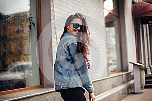 Happy attractive young woman in sunglasses walking in the city. Playful woman wraps herself over her shoulder and looking in