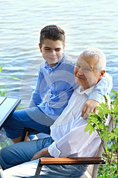 Happy Ñaucasian teen boy and elderly grandfather in blue shirts and jeans sitting by the lake. Relaxing outdoors together. Family