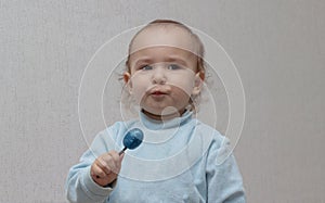 Happy baby in a blue sweater eating a blue Lollipop candy, on a light background. Beautiful little girl with brown eyes with a