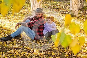 Happy baby boy and bearded dad in hat sitting on fallen yellow autumn leaves. Father and son walk in the parkand having