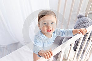 A happy baby boy in a crib in the children`s room is holding on to the side and smiling