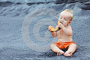 Happy baby boy eating orange papaya fruit on black beach