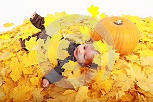 Happy baby boy lying by pumpkin covered by maple leaves