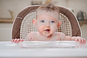 Happy baby with food stained mouth sitting on child chair, home kitchen background