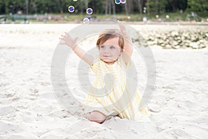 happy baby girl on beach on sand in striped yellow summer dress plays with soap bubbles on sunny day