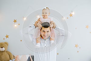 Happy baby one-year-old girl sits on dad`s shoulders and they smile in the children`s room