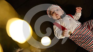 A happy baby in a red Santa Claus hat is lying on the floor, smiling happily and laughing on a dark background