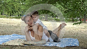 Happy baby sitting on the back of his mother, who is lying on the grass, laughing and hugging her mother`s neck. Mom and