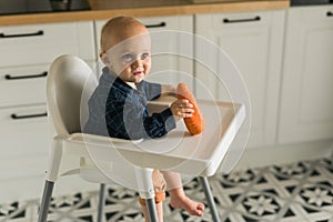 Happy baby sitting in high chair eating carrot in kitchen copy space. Healthy nutrition for kids. Bio carrot as first