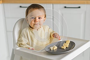 Happy baby sitting in high chair eating fruit in kitchen. Healthy nutrition for kids. Bio carrot as first solid food for