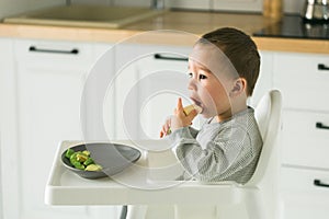 Happy baby sitting in high chair eating fruit in kitchen. Healthy nutrition for kids. Bio carrot as first solid food for