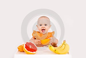 Happy baby sitting in high chair eating fruits in a white kitchen. Healthy nutrition for kids. Children eat oranges and