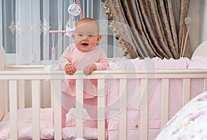 Happy baby standing in crib in pink bedroom scene.