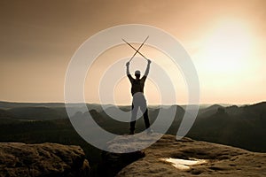 Happy backpacker with x crossed poles in the air, open misty mountain valley bellow cliff. Silhouette of tourist