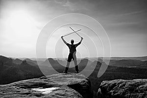 Happy backpacker with x crossed poles in the air, open misty mountain valley bellow cliff. Silhouette of tourist