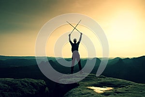 Happy backpacker with x crossed poles in the air, open misty mountain valley bellow cliff. Silhouette of tourist