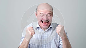 Happy bald senior man in shirt celebrating his success over gray background.