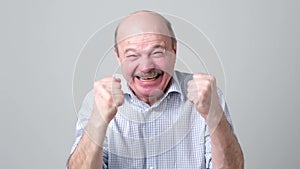 Happy bald senior man in shirt celebrating his success over gray background.