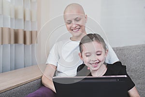 Happy bald woman with alopecia smiles and joyfully spends time with her child watching cartoons on a tablet