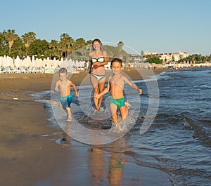 Happy barefoot family having fun - mother, son, together run with splashes by water pool along surf of sunset sea beach. Active