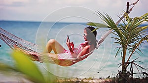 Happy beach woman wearing sunglasses reading sms texting on smartphone lying on hammock relaxing on tropical vacation