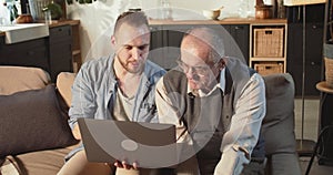 Happy bearded 30s young adult man helping senior retired 70s dad use laptop and internet on couch at home living room.