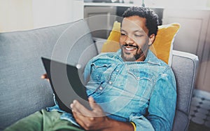 Happy bearded African man spending free time in sofa and using digital tablet at modern home.Concept of young people