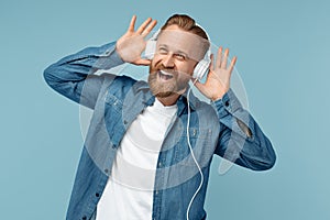 Happy bearded blonde hipster man in jeans shirt listening music in white stylish headphone over blue background.