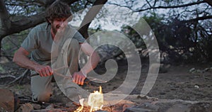 Happy bearded caucasian male survivalist adding kindling to fire at camp in wilderness