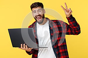 Happy bearded hipster young man showing peace gesture on notebook or computer over yellow background. Open mouth.