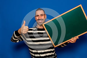 Happy bearded hispanic man showing, presenting and holding blank white board with thumb up isolated over blue background