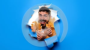 Happy bearded man with autumn leaves looking through paper hole. Smiling man in trendy autumn outfit with fall maple