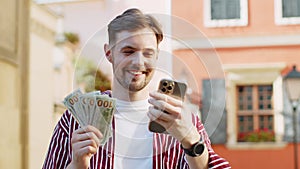 Happy bearded man counting money dollar cash, use smartphone calculator app in urban city street