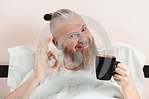 Happy bearded man enjoying morning coffee in bed showing OK gesture. Smiling hipster guy starting the day. Closeup