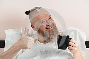 Happy bearded man enjoying morning coffee in bed showing thumb up gesture. Smiling hipster guy starting the day. Closeup