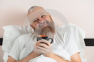 Happy bearded man enjoying morning coffee in bed. Smiling hipster guy starting the day. Closeup