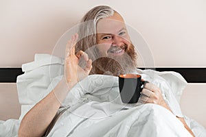 Happy bearded man enjoying morning coffee in cozy bed showing ok sign. Smiling guy looking at camera. Closeup