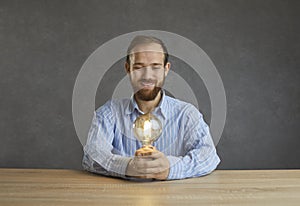 Happy bearded man holding a yellow light bulb sitting at the table on a gray studio background.