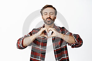 Happy bearded man, husband shows heart sign and kissing, pucker lips with closed eyes, standing over white background