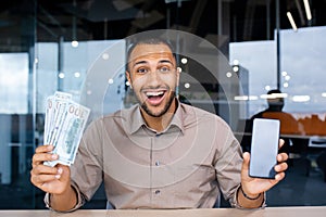 Happy bearded man in office holding stack of dollars and showing phone screen to camera, happy with winning, online
