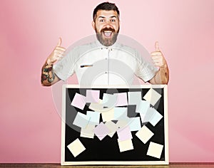 Happy bearded man standing at table with sticky notes on blackboard shows thumbs up. Notes, reminder.