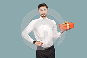 Happy bearded man in white shirt standing with hand on waist holding red gift box, looking at camera with satisfied face and