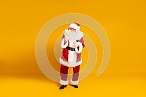 Happy bearded real Santa Claus with headphones dancing and listening music from mobile phone. Yellow studio background. Christmas