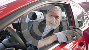 Happy bearded senior man showing thumb up and smiling at camera sitting on driver's seat in pricey new automobile in