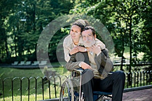 Happy bearded senior man in wheelchair with charming young girl supporting disabled grandparent on walking in park