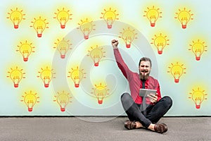 Happy bearded young businessman in red shirt holding tablet sitting on floor, celebrating amazing idea or victory, looking at